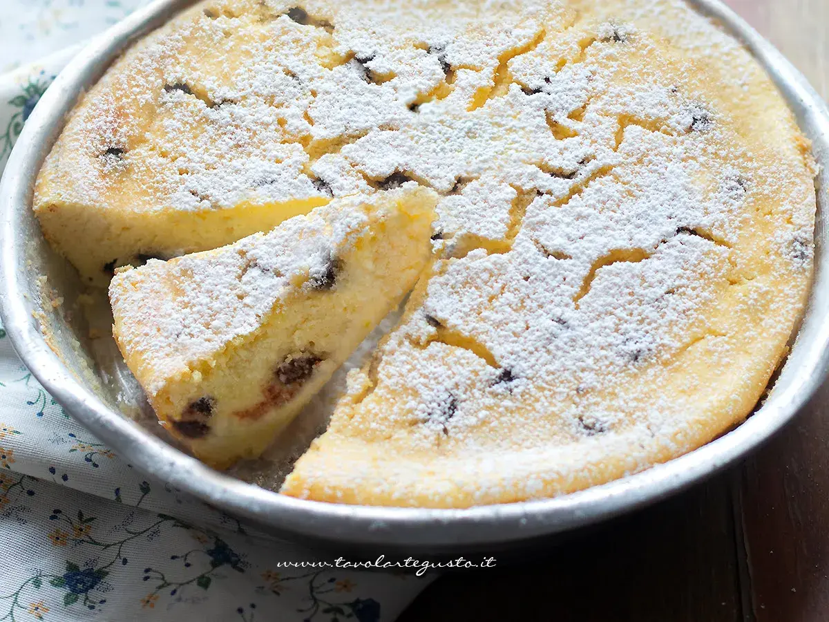 Torta soffice ricotta e cioccolato, cosparsa di zucchero a velo, in una tortiera. Una fetta è già stata tagliata.