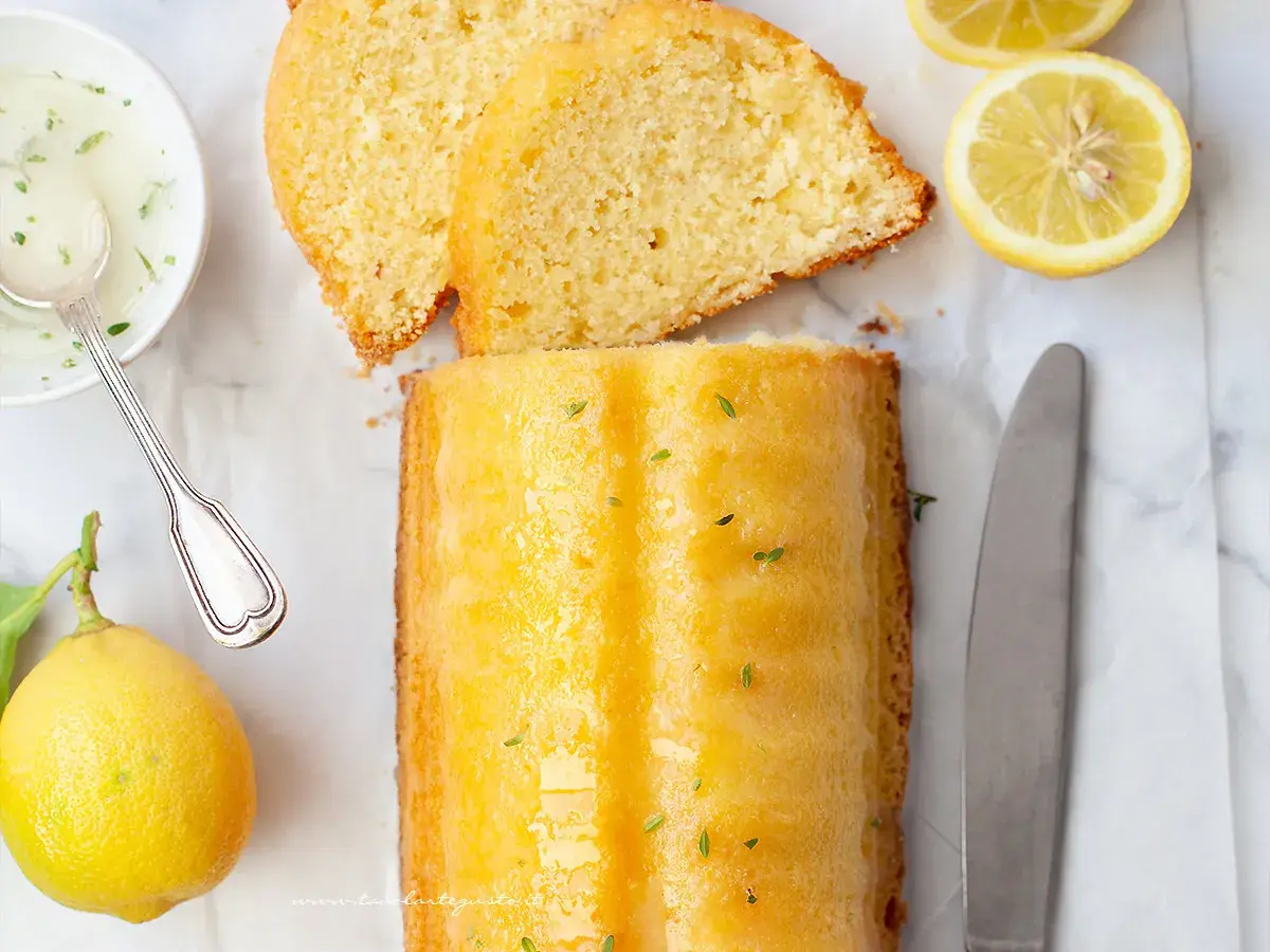 Un delizioso plumcake al limone glassato, con fette affettate, limoni freschi, una ciotolina di glassa e un coltello.