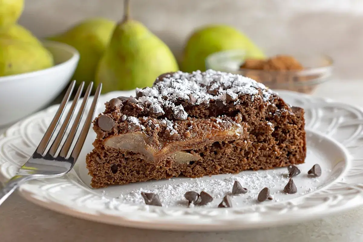 Una fetta di torta pere e gocce di cioccolato, cosparsa di zucchero a velo e gocce di cioccolato, su un piatto bianco con una forchetta.