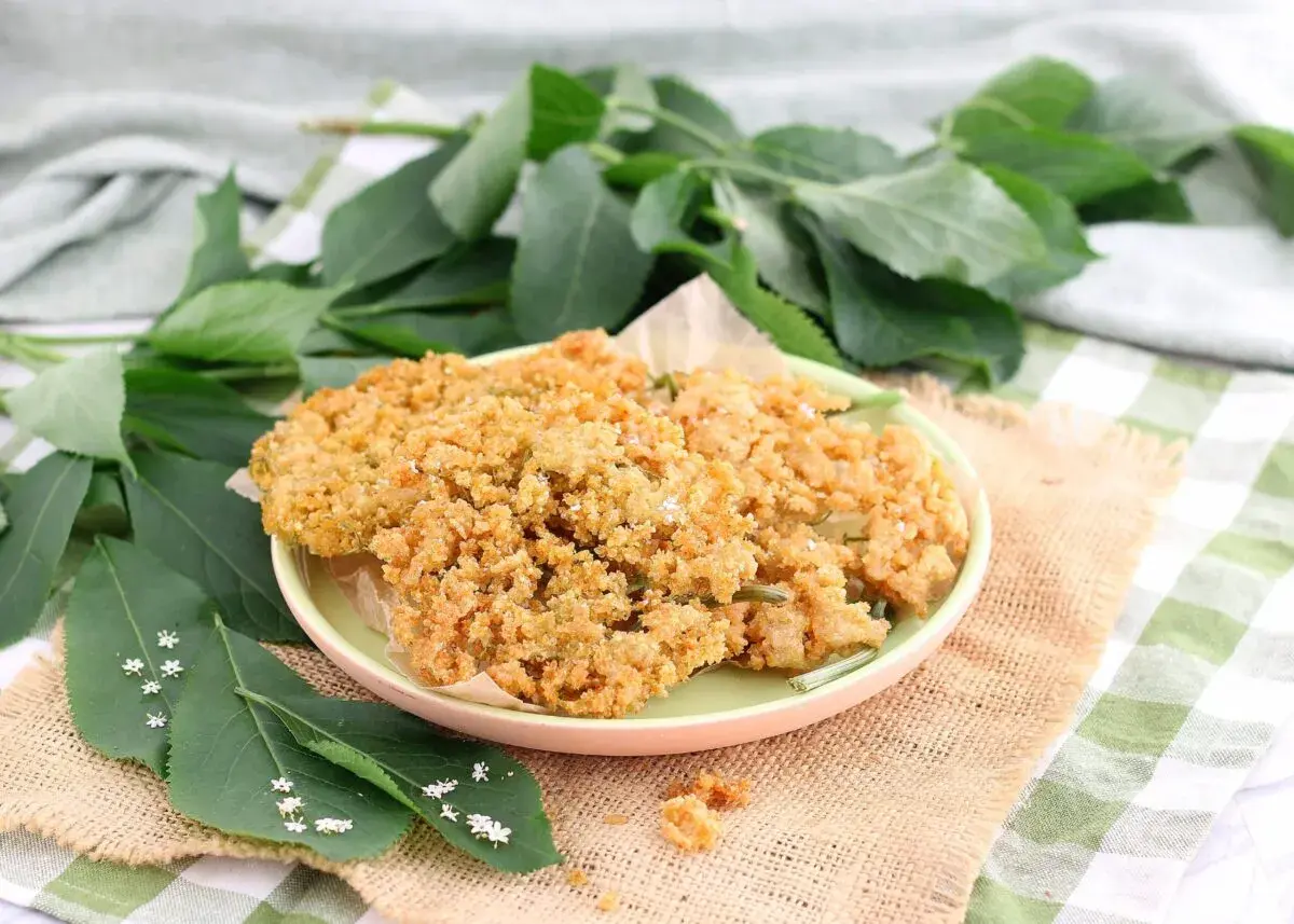 Fiori di sambuco fritti dorati e croccanti su un piatto, con foglie verdi e piccoli fiori di sambuco tutt'intorno.