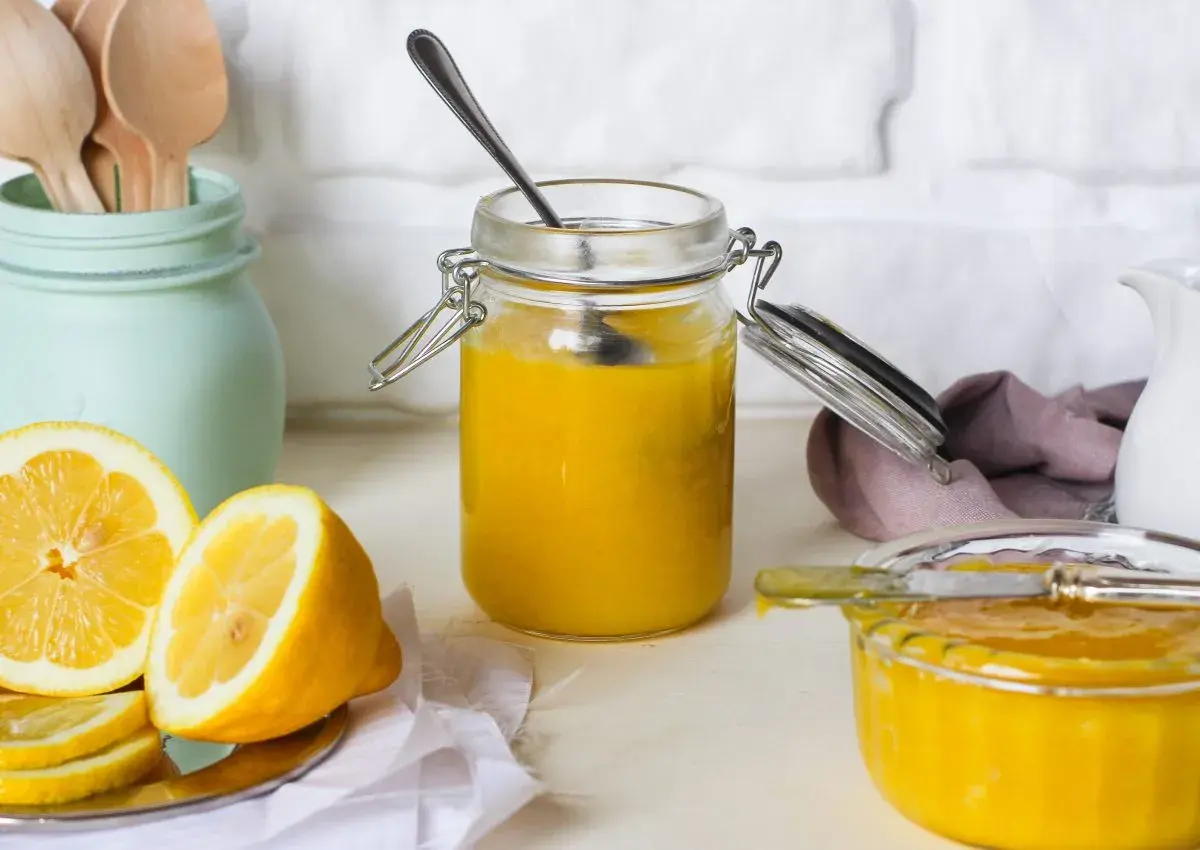 Vasetto di vetro pieno di lemon curd, con limoni freschi affettati e un cucchiaino.