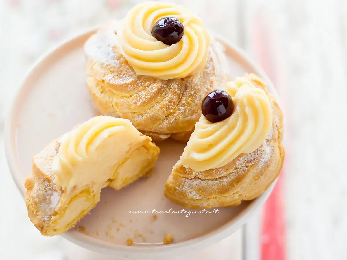 Zeppole di San Giuseppe, dolci soffici con crema pasticcera e ciliegina, uno è tagliato per mostrarne l'interno goloso.