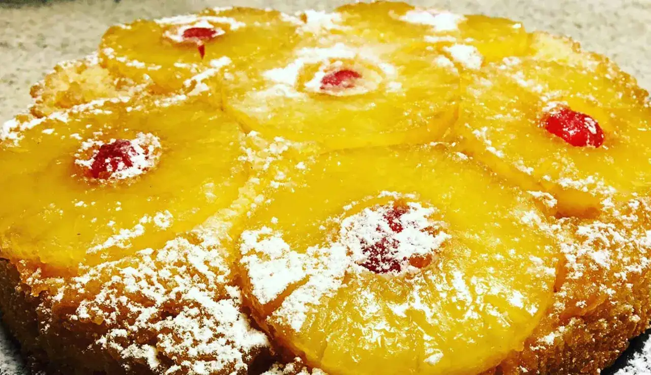 Torta ananas soffice e dorata, decorata con fette di ananas caramellato e ciliegie rosse, spolverata di zucchero a velo.