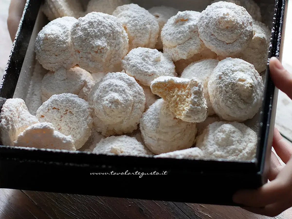 Scatola piena di dolci di cocco soffici e spolverati di zucchero a velo. Uno è spezzato, rivelando un interno morbido e invitante.