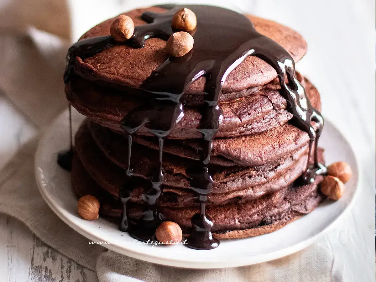 Pancake cacao scuri e soffici, inzuppati di salsa al cioccolato e guarniti con nocciole intere.