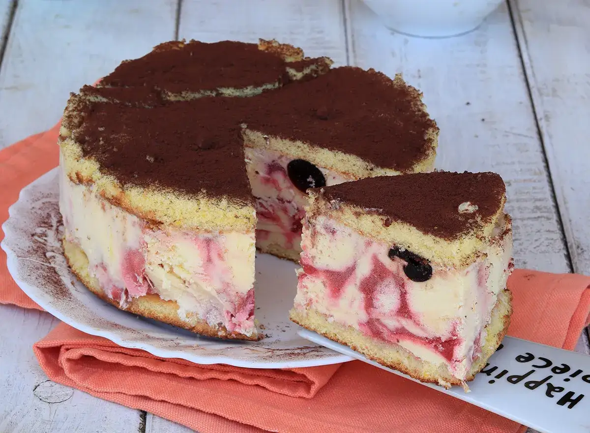 Deliziosa torta gelato con pan di Spagna, crema e ciliegie, spolverata di cacao. Una fetta &egrave; gi&agrave; servita su un piattino.