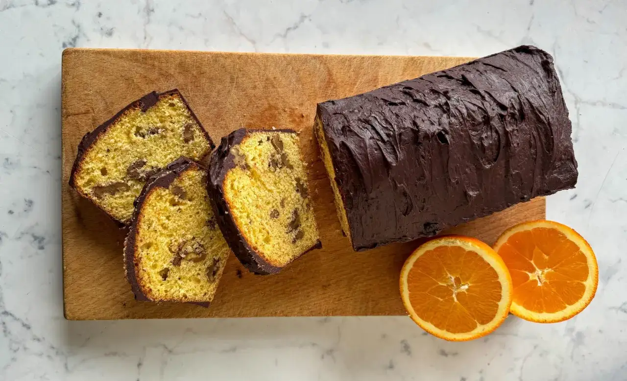 Plumcake noci e cioccolato, soffice e goloso, con glassatura scura e fette che rivelano il suo interno ricco. Accompagnato da spicchi d'arancia.