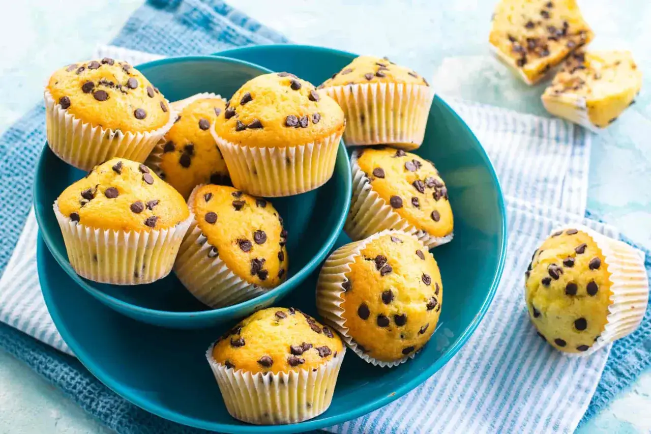Muffin senza uova con gocce di cioccolato, dorati e invitanti, serviti in una ciotola blu su un tovagliolo a righe.