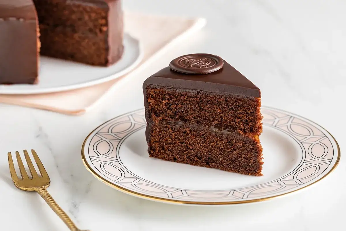 Una fetta di torta al cioccolato, ricoperta di glassa lucida e decorata con un sigillo di cioccolato. Un'altra torta al cioccolato è visibile sullo sfondo.