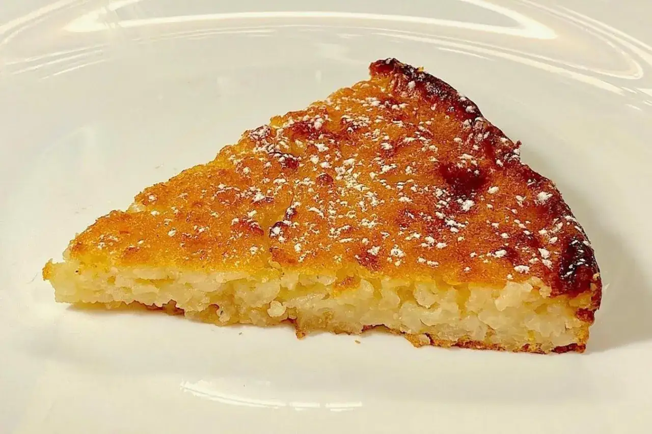 Una fetta di torta di riso massese, dorata e cosparsa di zucchero a velo, su un piatto bianco.