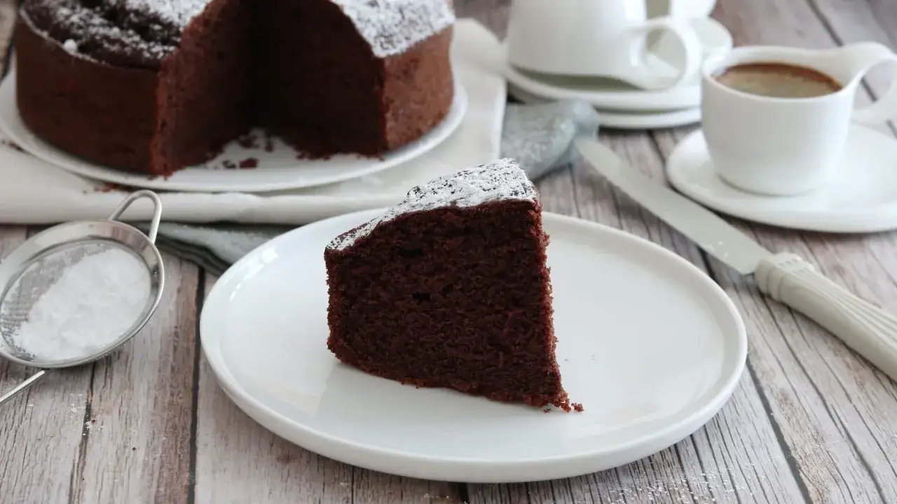 Una fetta di torta al cioccolato soffice, preparata con albumi, cosparsa di zucchero a velo. Accanto, una tazza di caffè.