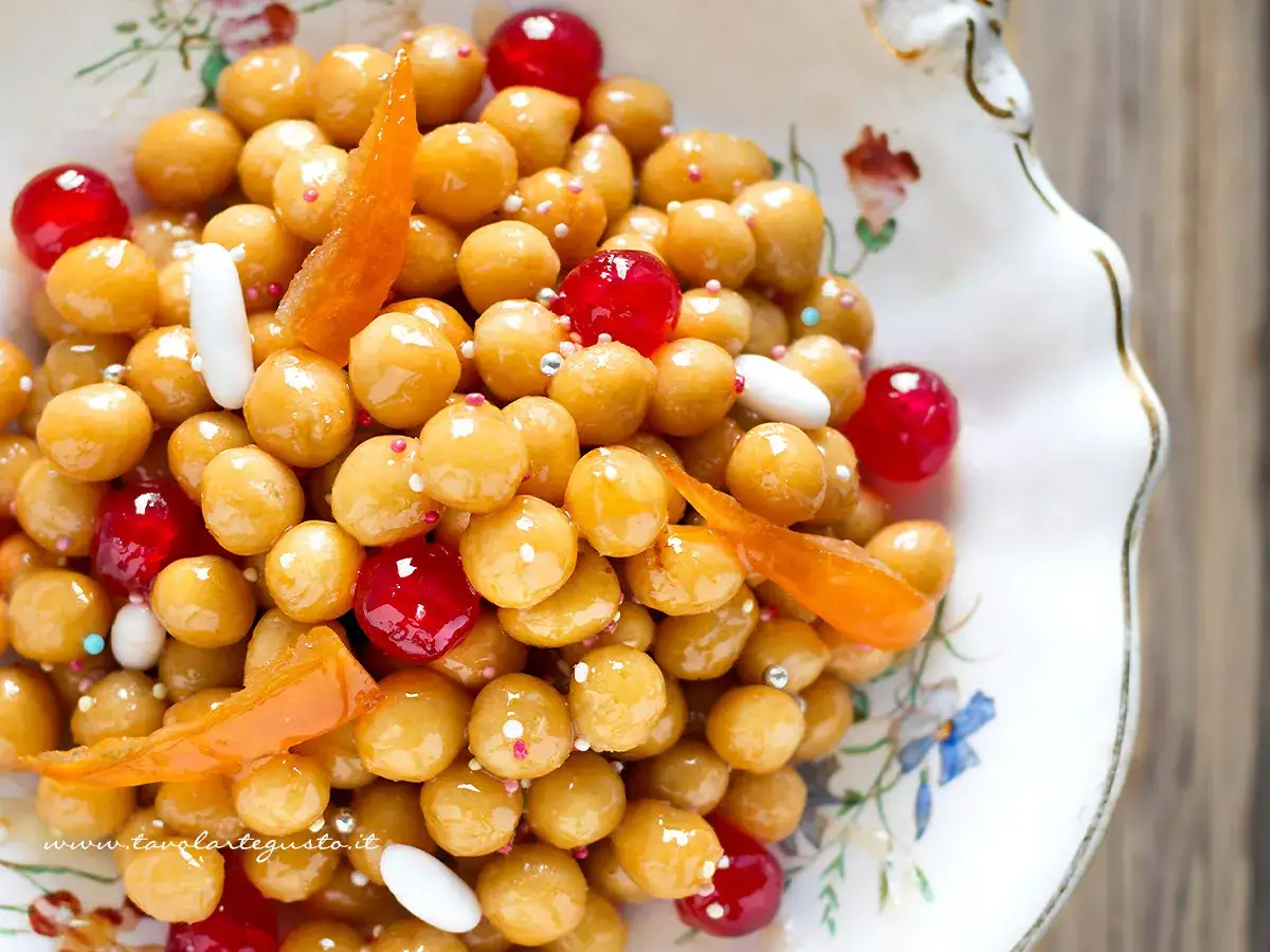 Struffoli ricetta napoletana: palline dorate glassate, ciliegie rosse, scorzette d'arancia e confettini in un piatto decorato.