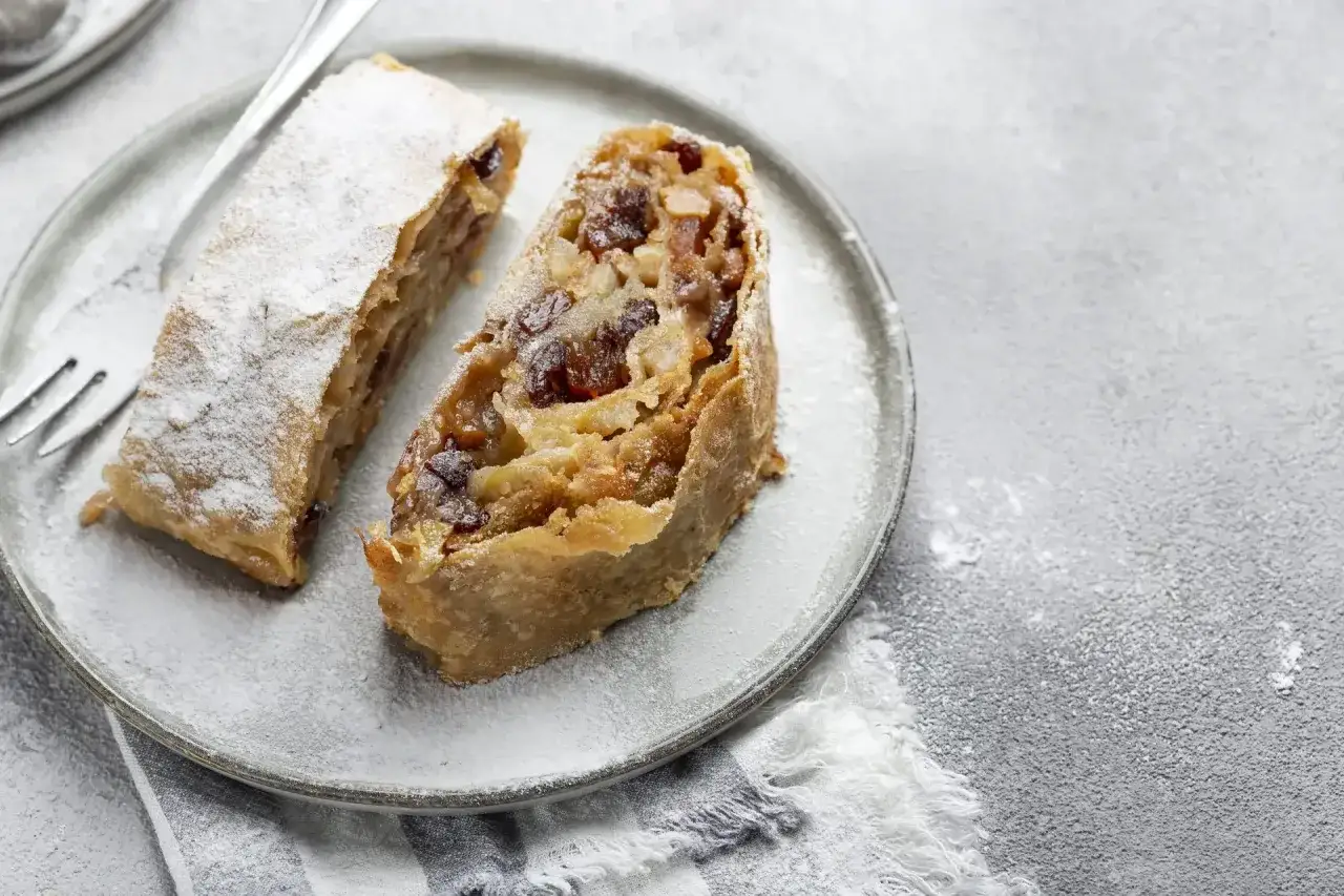 Due fette di strudel di mele su un piatto, cosparse di zucchero a velo. La pasta sfoglia dorata racchiude un ripieno ricco di mele e uvetta.