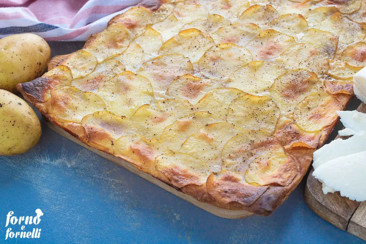 Torta salata con patate su pasta sfoglia dorata, pronta per la cottura. Accanto, patate fresche e formaggio.