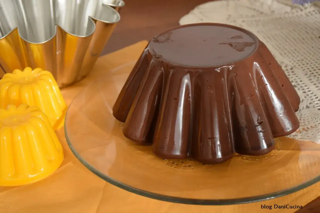 Budino al cioccolato lucido e invitante, pronto per essere gustato. Accanto, due piccoli budini gialli e uno stampo metallico.