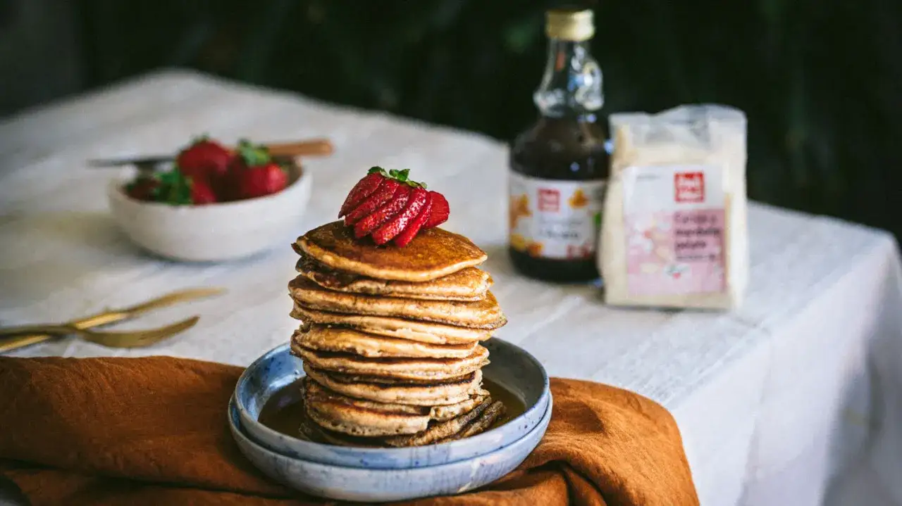 Pila di pancake con farina di mandorle, guarniti con fragole fresche, su un piatto blu. Sullo sfondo, una ciotola di fragole, una bottiglia di sciroppo e un sacchetto di farina.