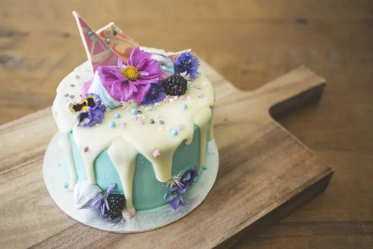 Torta decorata con glassa fondente bianca, fiori viola, more e meringhe su un tagliere di legno.