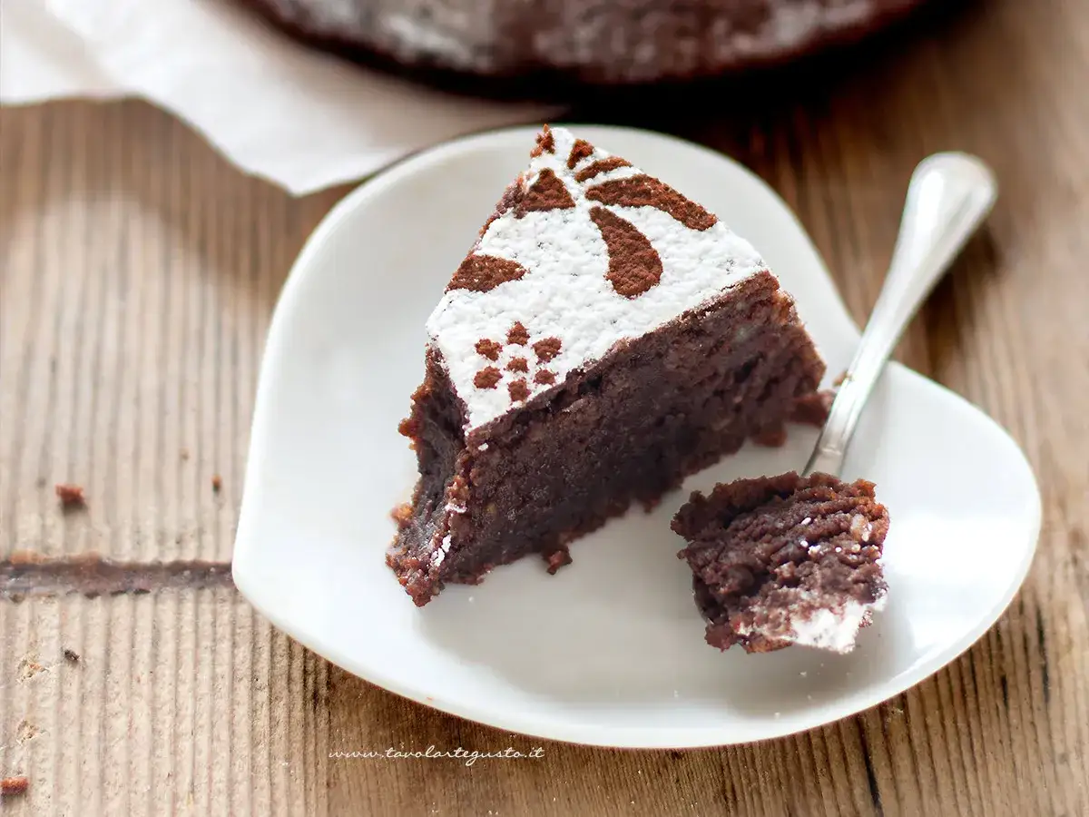 Una fetta di torta caprese ricetta facile, cosparsa di zucchero a velo e cacao, su un piatto bianco a forma di cuore. Un cucchiaino ha prelevato un pezzetto.