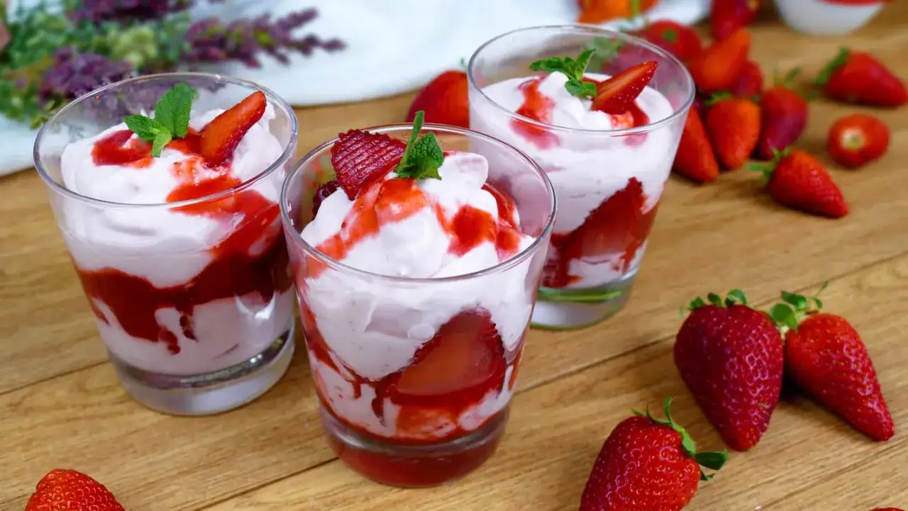 Delizioso mousse di fragole in bicchieri trasparenti, guarnito con fragole fresche e menta. Intorno, fragole rosse brillanti su un tavolo di legno.