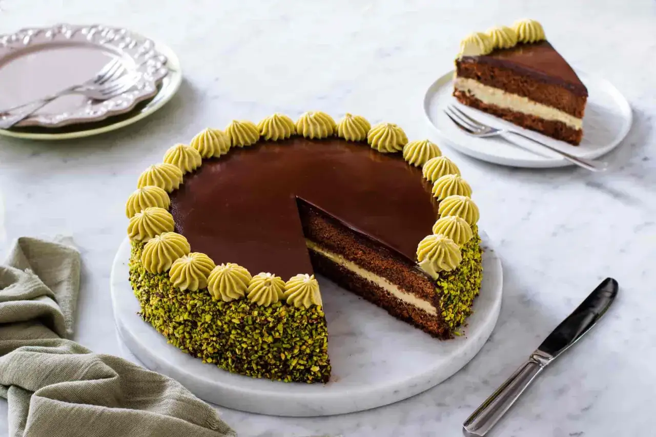 Torta al cioccolato con glassa lucida, bordi ricoperti di pistacchio tritato e decorazioni di crema al pistacchio. Una fetta è già stata servita.