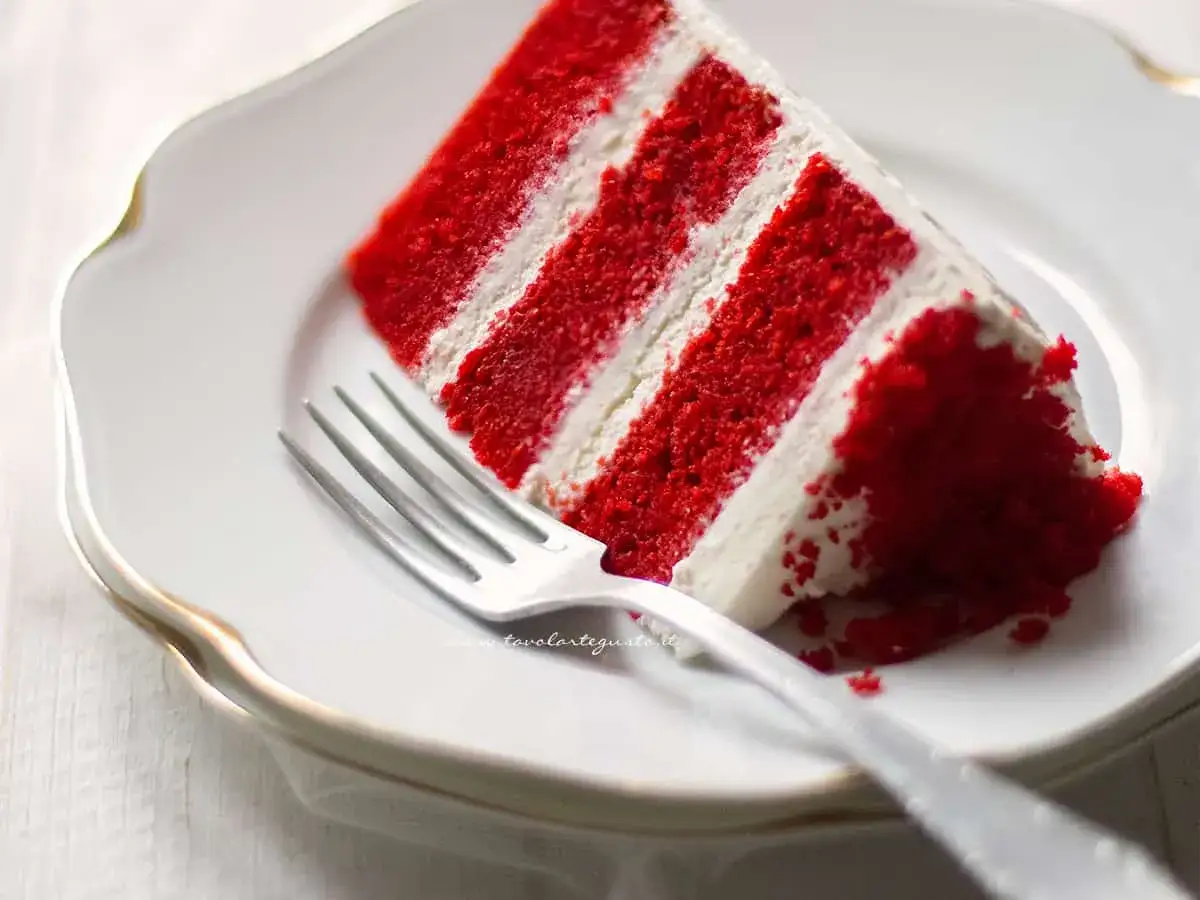Una fetta di torta red velvet su un piatto bianco, con una forchetta. La torta ha strati rossi e crema bianca.