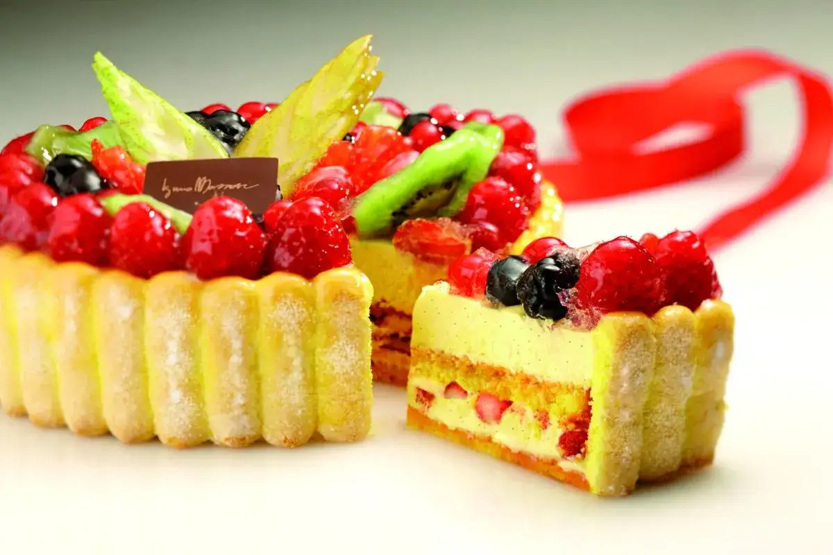 Una torta charlotte riccamente decorata con frutti di bosco freschi, kiwi e fettine di pera, con un nastro rosso sullo sfondo.