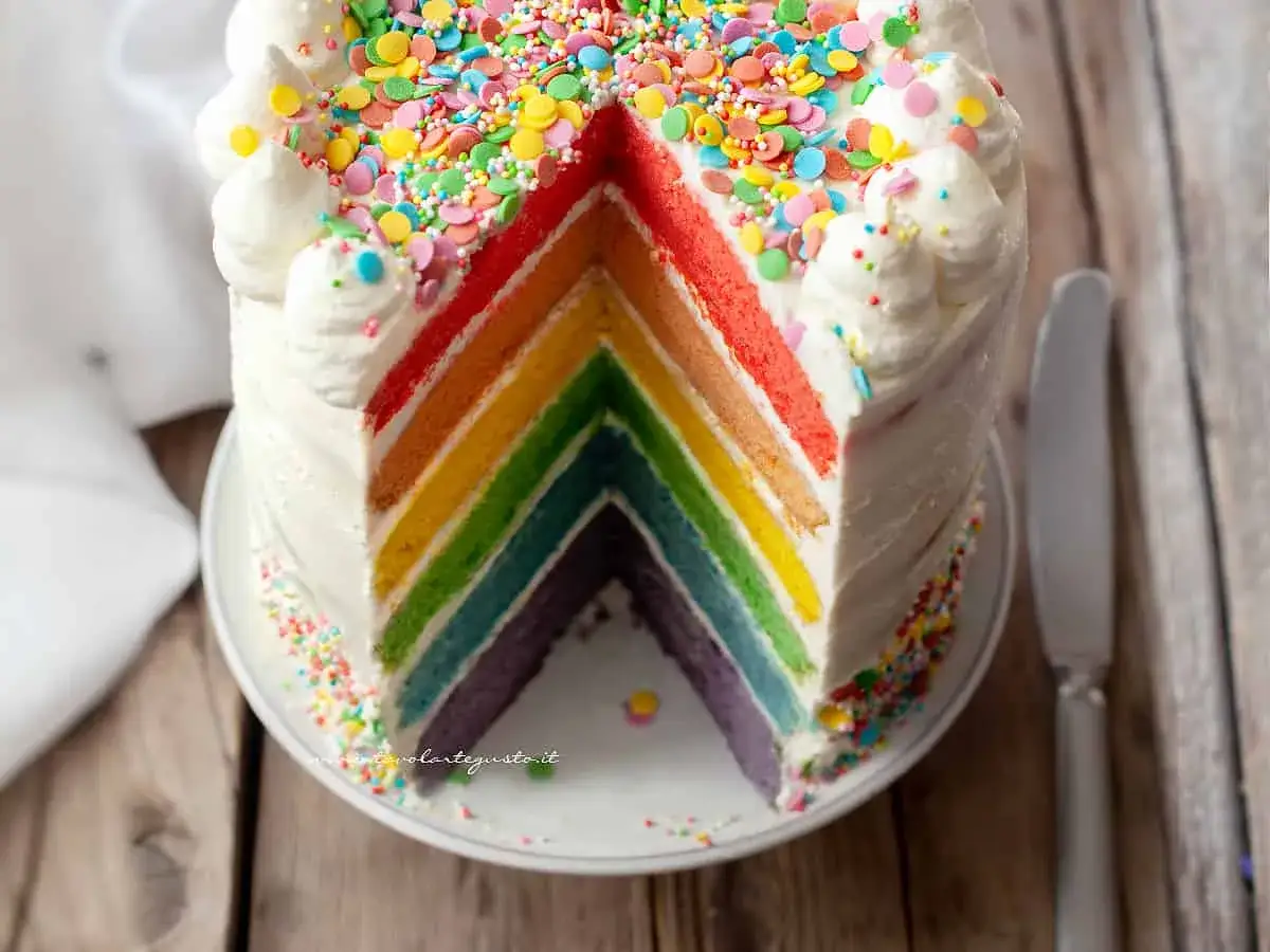 Una fetta di torta arcobaleno, con strati colorati e glassa bianca decorata con zuccherini.