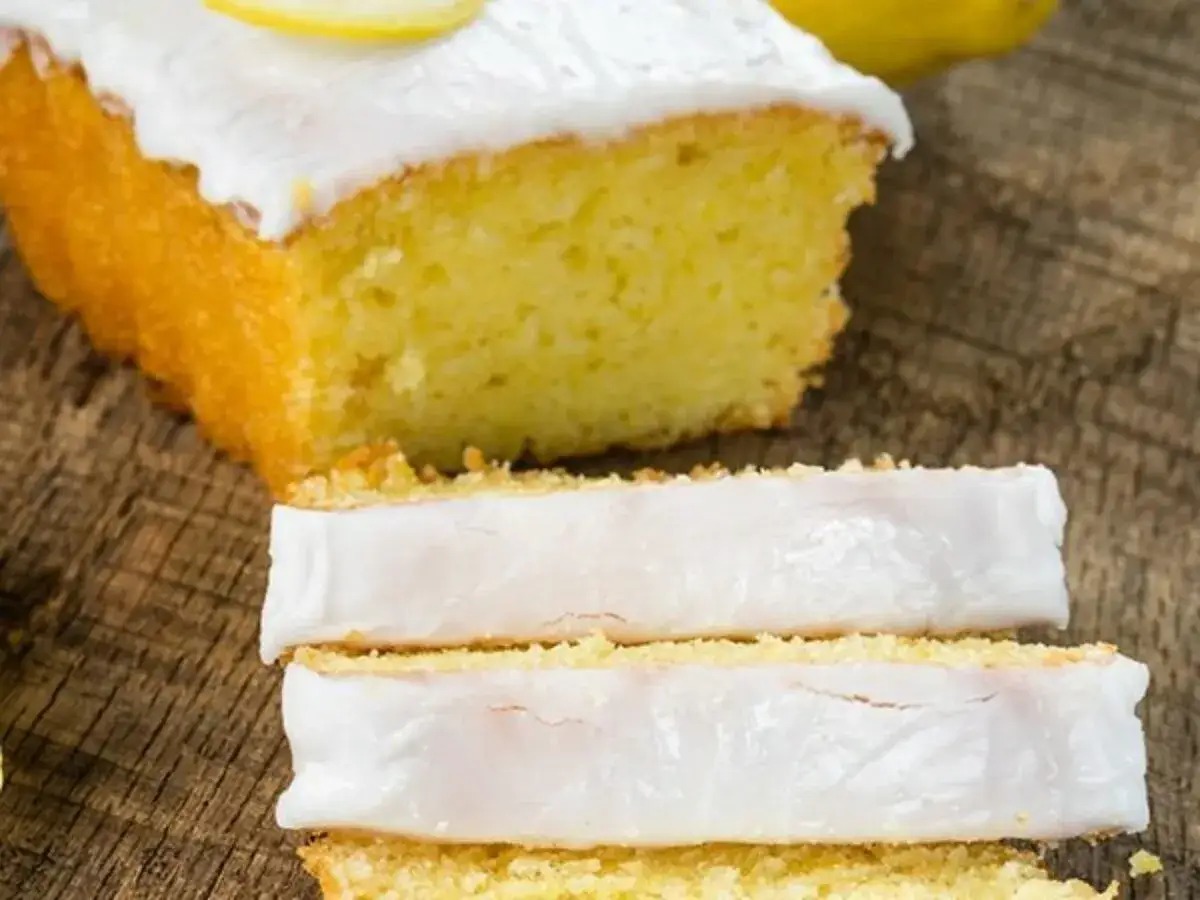 Fetta di plumcake al limone con glassa bianca e una fetta di limone in cima, su un tagliere di legno.