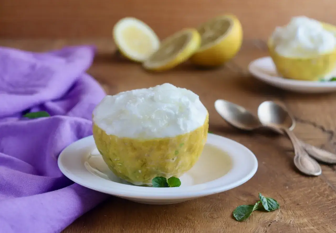 Sorbetto al limone senza albume servito in un limone scavato, con limoni affettati e un tovagliolo viola sullo sfondo.