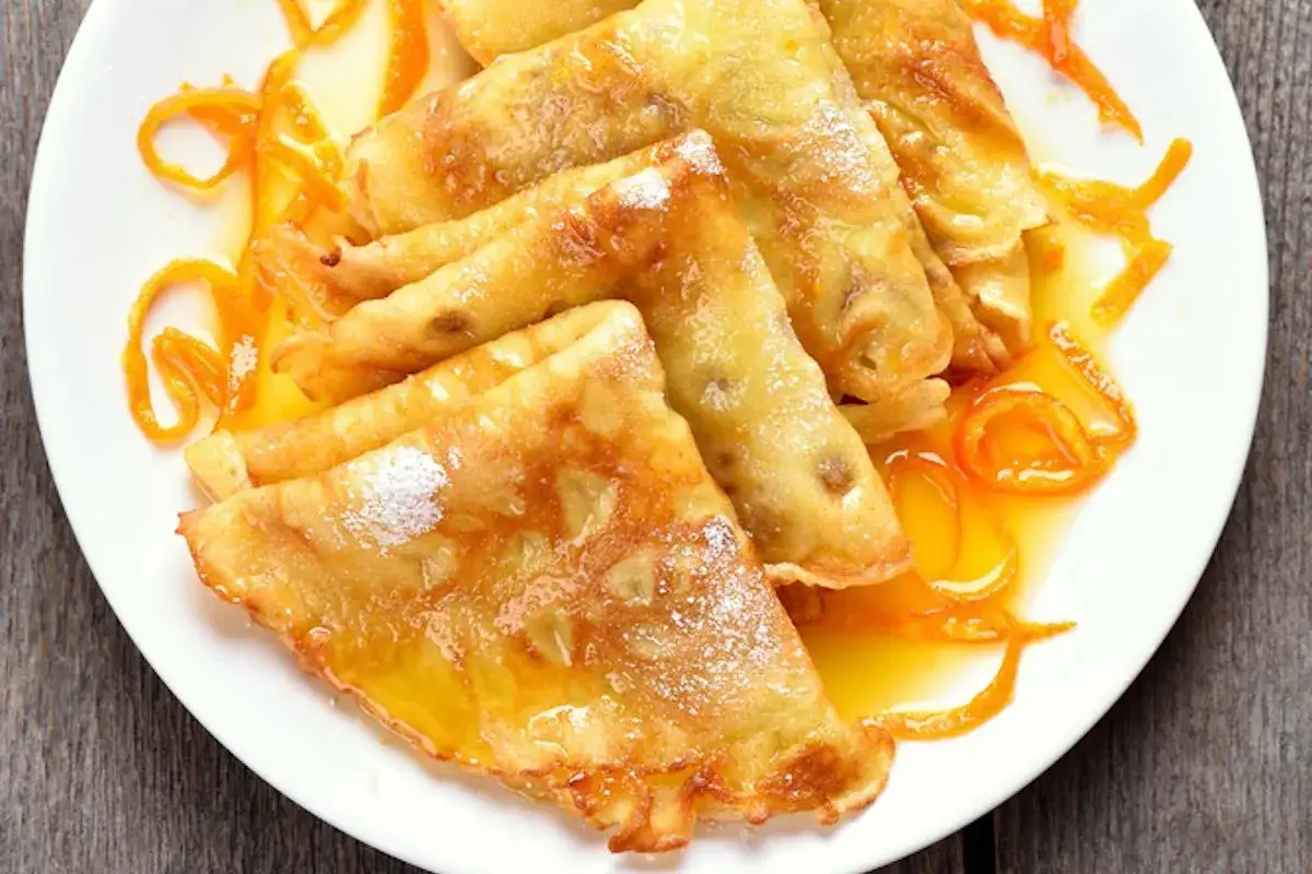 Crepes Suzette dorate e lucide, cosparse di zucchero a velo e immerse in una salsa all'arancia con scorzette candite.