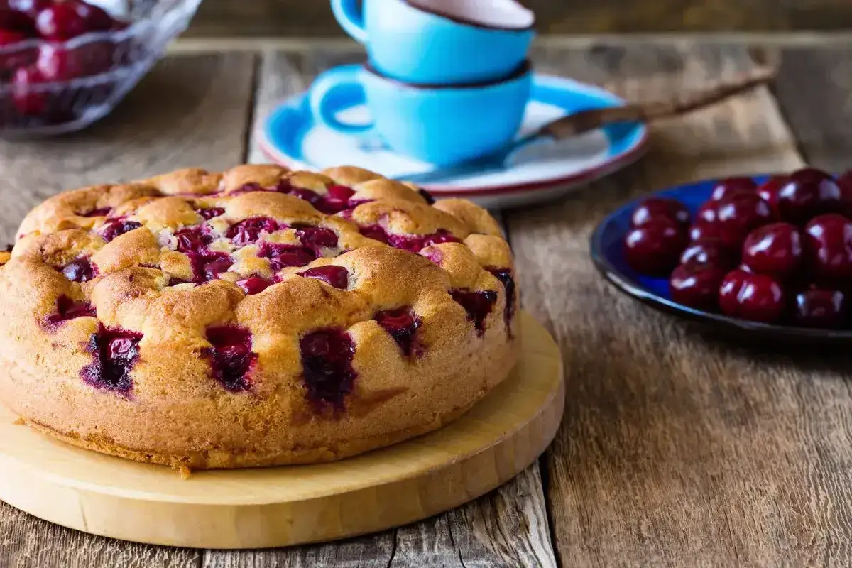 Torta con ciliegie dorata, con ciliegie rosse succose incastonate nell'impasto. Accanto, una ciotola di ciliegie fresche e tazze blu.