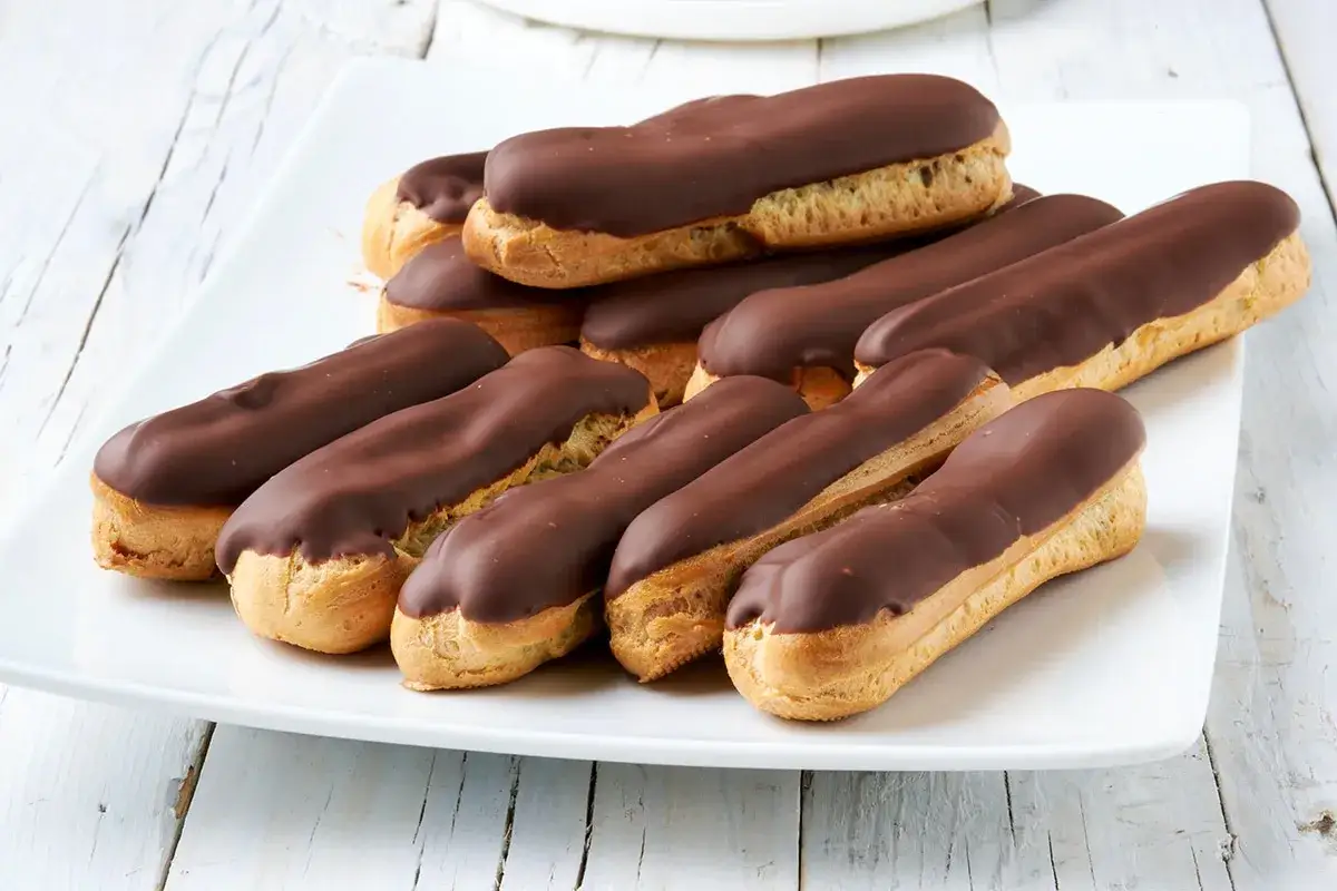 Un piatto bianco pieno di dolci eclair ricoperti da lucido cioccolato fondente, pronti per essere gustati.
