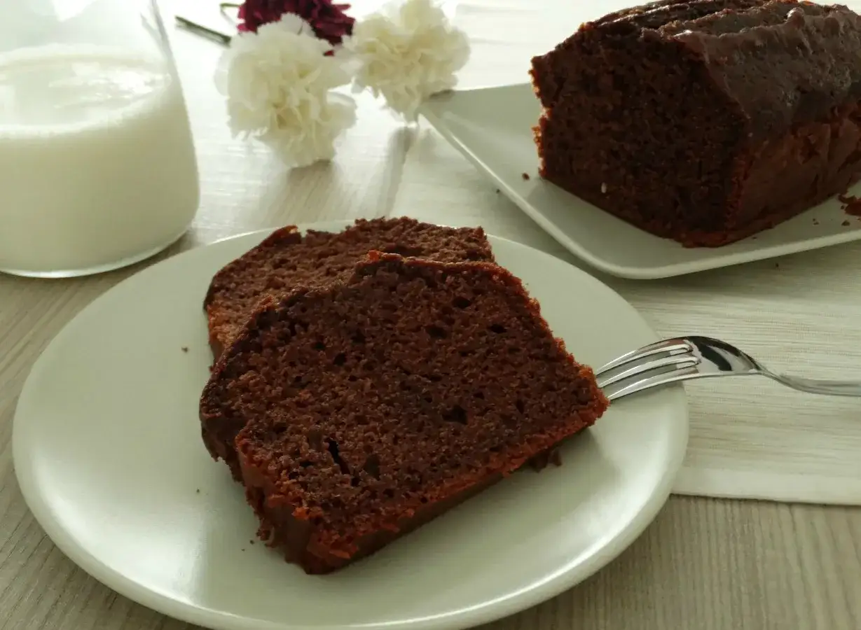 Fette di plumcake yogurt e cioccolato su un piatto, con un bicchiere di latte e fiori sullo sfondo.