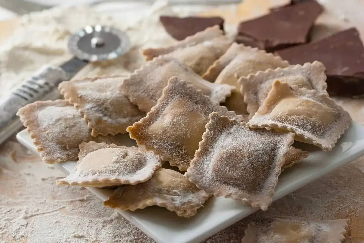 Tortelli al cioccolato appena fatti, spolverati di zucchero a velo, pronti per essere gustati. Sullo sfondo, pezzi di cioccolato fondente e un rotella tagliapasta.