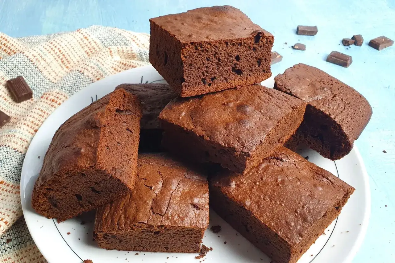 Un mucchio di deliziosi brownies senza glutine, tagliati a cubetti e disposti su un piatto bianco, con qualche pezzetto di cioccolato sparso intorno.