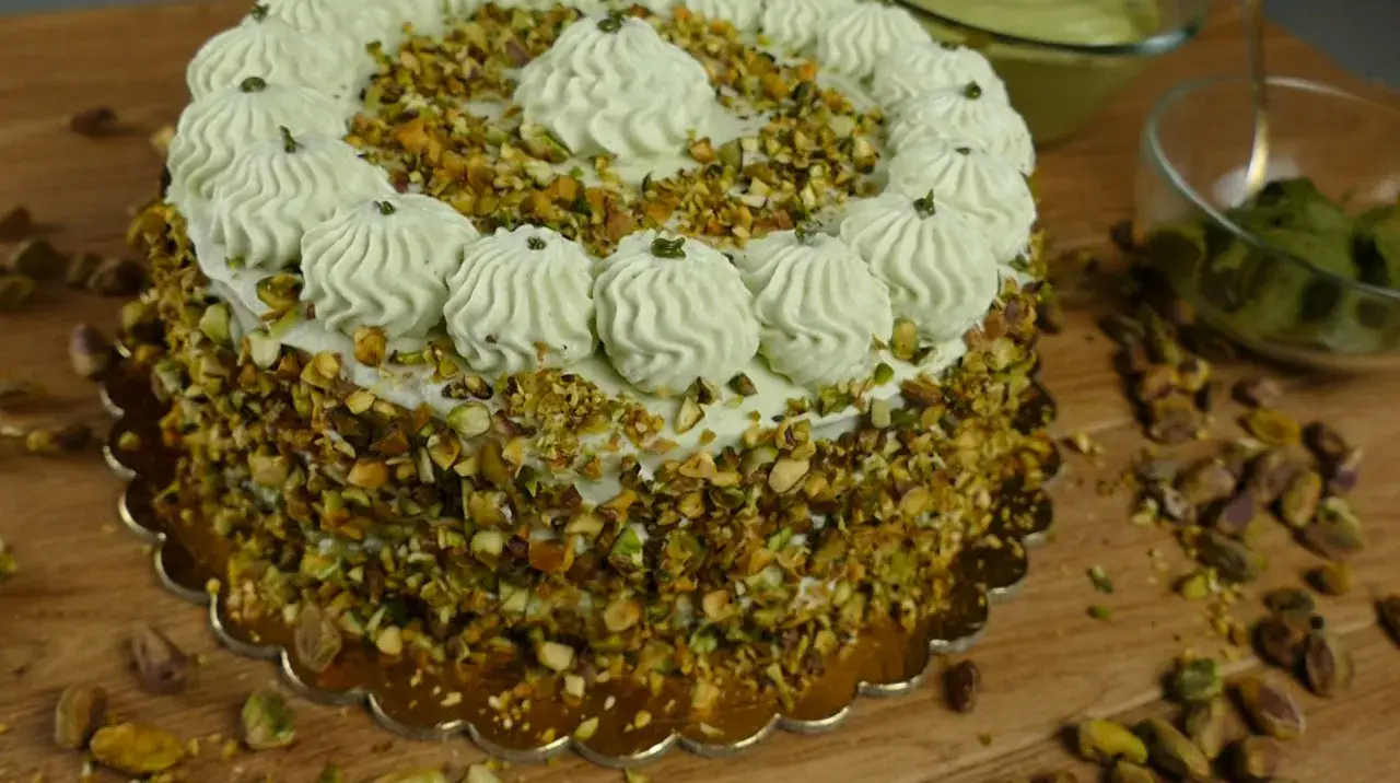 Una deliziosa torta al pistacchio, ricoperta di crema e granella di pistacchi, con decorazioni a ciuffetti e pistacchi sparsi intorno.