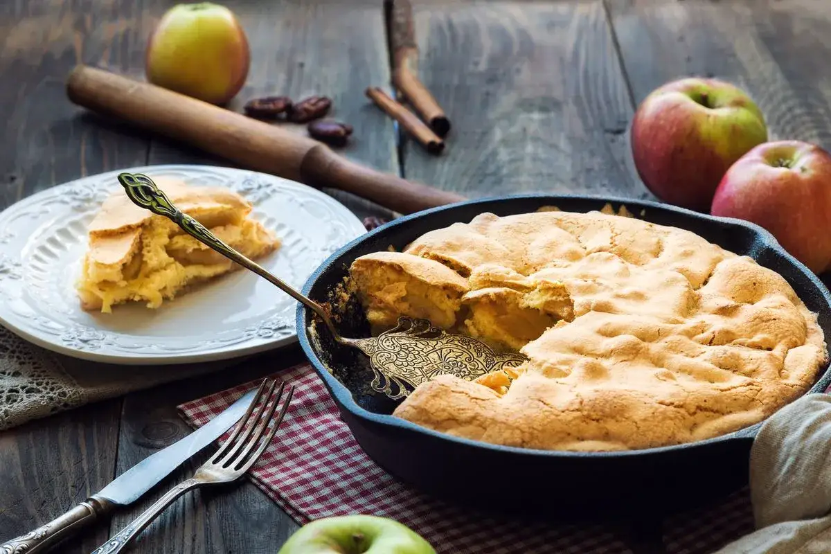 Una deliziosa torta di mele in padella, appena sfornata, con una fetta servita su un piatto. Mele rosse e verdi sullo sfondo.