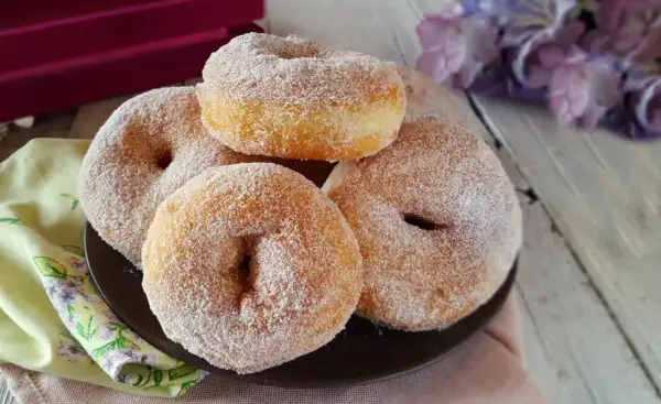 Graffe senza patate, soffici e dorate, ricoperte di zucchero. Un dolce invitante su un piatto scuro, con fiori viola sullo sfondo.