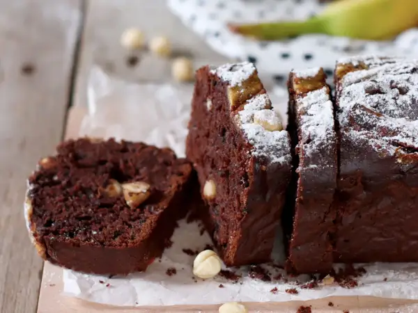 Fetta di banana bread cacao con nocciole e zucchero a velo, su tagliere di legno.