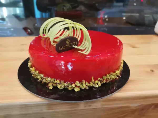 Torta con glassa a specchio lamponi, decorata con riccioli di cioccolato bianco e pistacchi tritati.