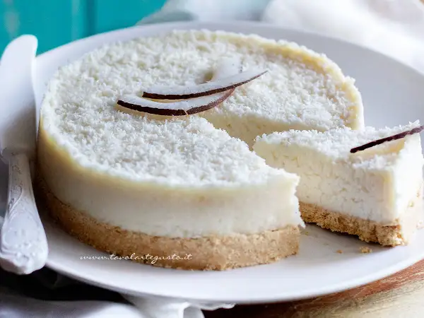 Delizioso cheesecake al cocco, ricoperto di cocco grattugiato e decorato con scaglie di cocco fresco. Una fetta è già stata tagliata.