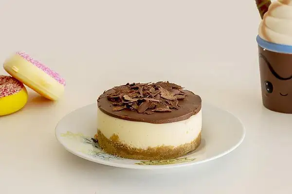 Mini cheesecake al cioccolato con base croccante e copertura di cioccolato fondente, accompagnato da due ciambelle colorate e una tazza sorridente.