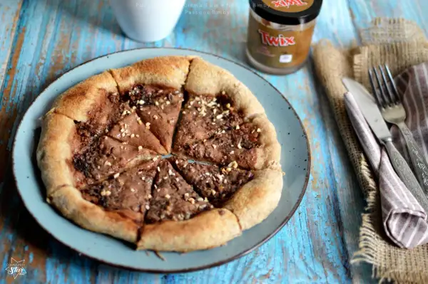 Pizza al cioccolato con crema Twix e nocciole, cosparsa di granella croccante. Un dolce goloso pronto da gustare.