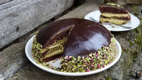 Torta al cioccolato con crema al pistacchio, ricoperta di glassa lucida e granella di pistacchio. Una fetta è già servita.
