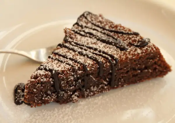 Una fetta di torta caprese al cioccolato, cosparsa di zucchero a velo e con una colatura di cioccolato fondente.
