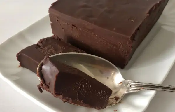 Una mattonella di cioccolato fondente, densa e lucida, su un piatto bianco. Un cucchiaino ne ha prelevato un pezzo.