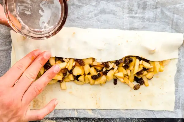 Mani che chiudono uno strudel di mele con pasta sfoglia, ripieno di mele a cubetti, uvetta e pinoli.