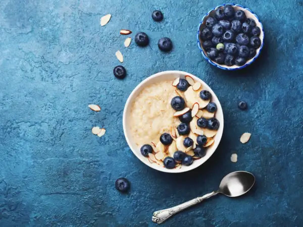 Porridge ricetta: una ciotola di avena con mirtilli, banane e mandorle a lamelle su sfondo blu.