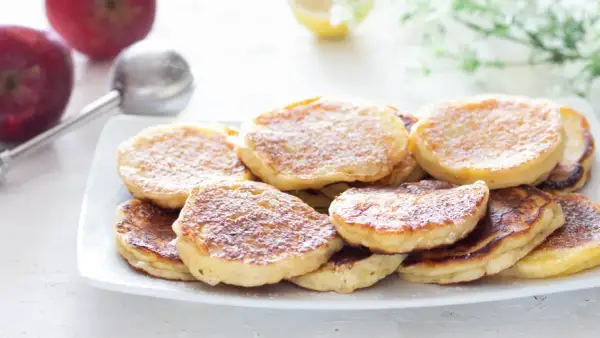 Piatto di frittelle di mele dorate e soffici, spolverate di zucchero a velo. Sullo sfondo, mele rosse e un colino.