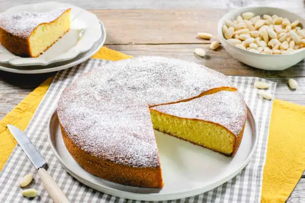 Deliziosa torta con farina di mandorle, spolverata di zucchero a velo, con una fetta tagliata. Accanto, una ciotola di mandorle.